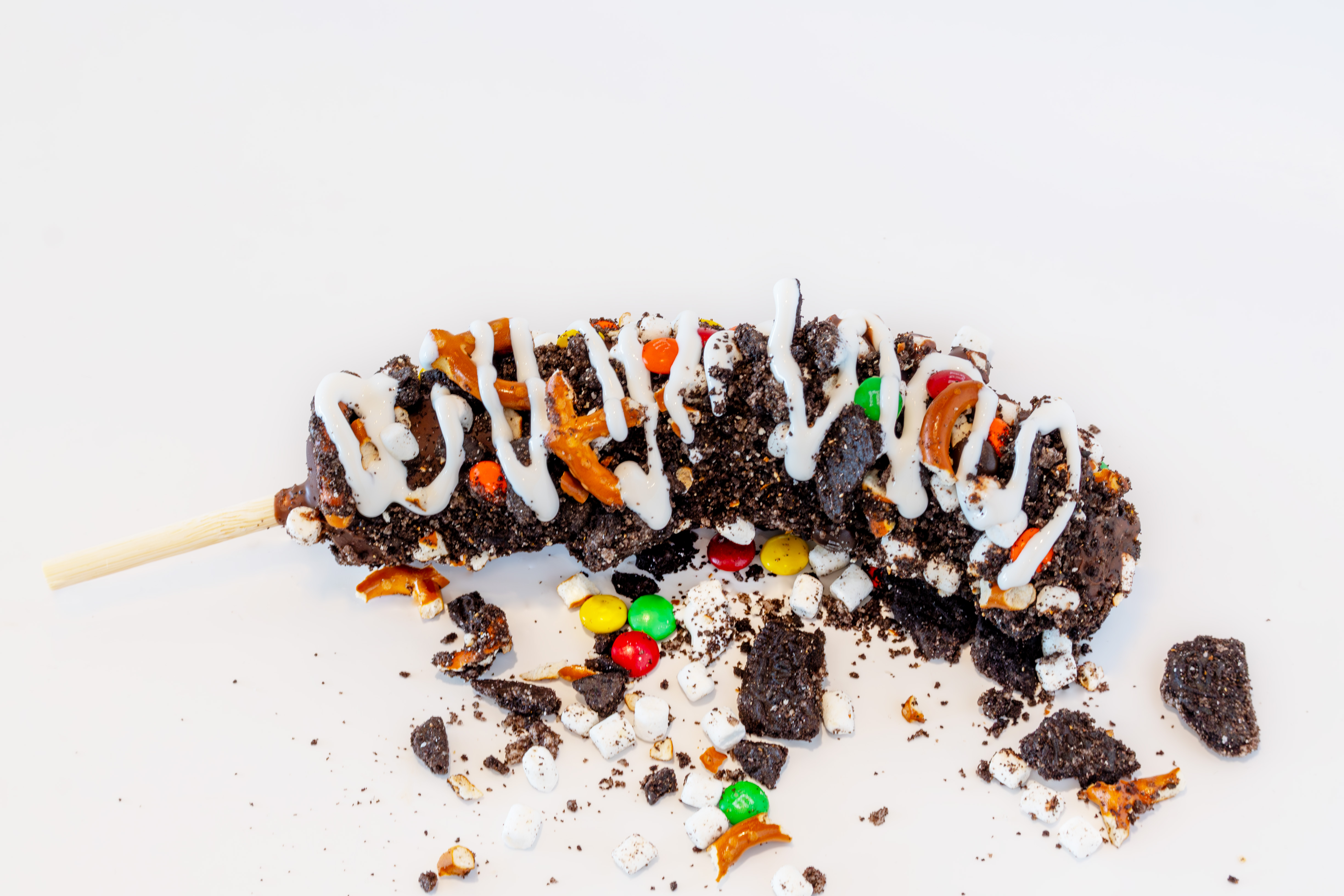 Overhead view of a chocolate dipped banana on a stick, covered with cookie crumbles, mini m&ms, and marshmallow drizzle
